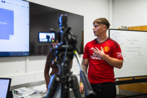 A young male student in a red Manchester United jersey giving a presentation on digital industries, with a camera and presentation screen in the background at Suffolk New College.