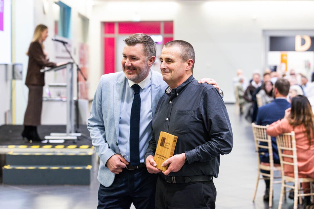 Two men smiling and shaking hands at Suffolk New College's annual dinner, celebrating reaching 200 industry partners, with a speaker at the podium and an audience in the background.