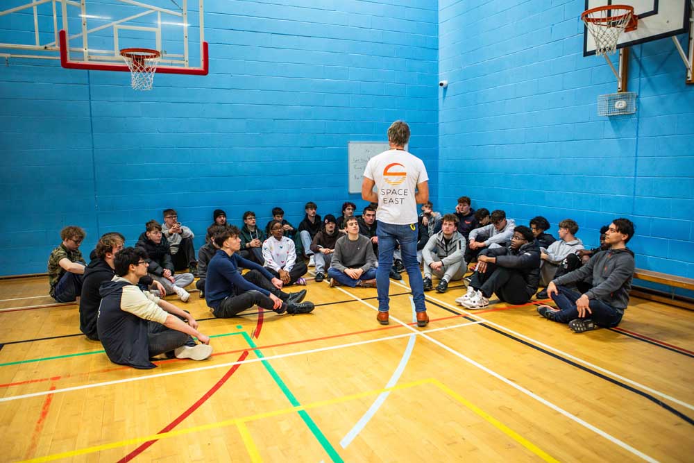 Suffolk New College students sitting on the gym floor listening to a speaker in a sports hall, promoting space exploration and STEM education.