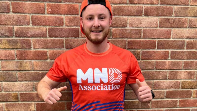An enthusiastic young man in a red MN Association t-shirt giving a thumbs-up against a brick wall, supporting MND fundraising efforts.