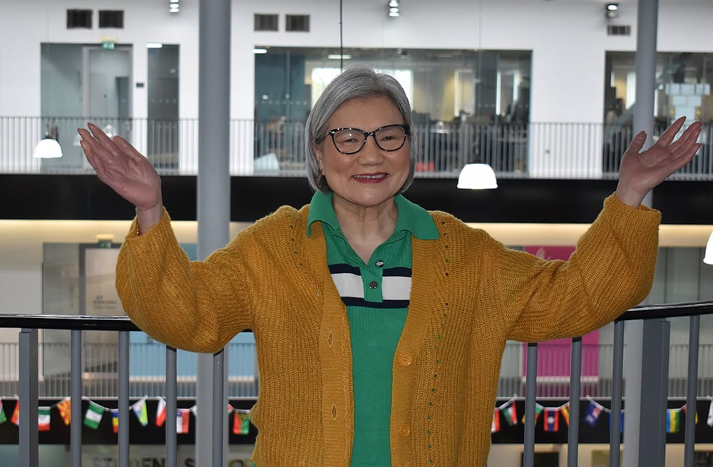An elderly woman with glasses, wearing a yellow cardigan and green shirt, smiling and raising her arms in a college atrium, highlighting lifelong learning and success at Suffolk New College.