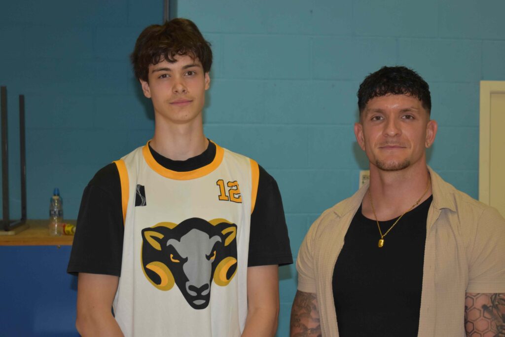 Two young men, one in a sports jersey and the other in casual clothing, standing indoors at Suffolk New College, showcasing student diversity and sports education.