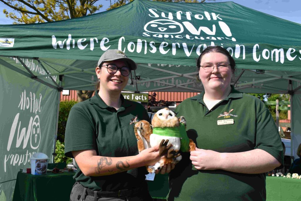 Two women representing Suffolk New College at the family fun day, holding a plush owl and a small animal, with a green Suffolk conservation tent in the background.