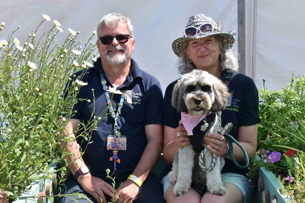 Two smiling adults and a dog sitting in a garden with flowers at Suffolk New College, showcasing a friendly and welcoming environment for students and staff.
