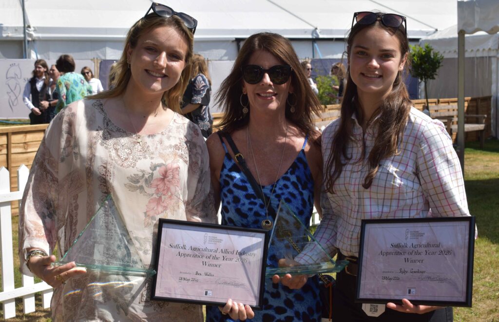 Three women at Suffolk New College outdoor event holding awards, smiling, with tents and other attendees in the background, celebrating achievement and success.