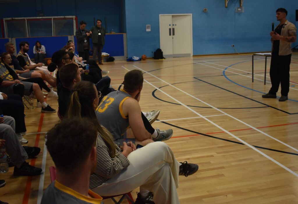 Students attending a sports event at Suffolk New College gymnasium, listening to a speaker during a college activity or presentation.