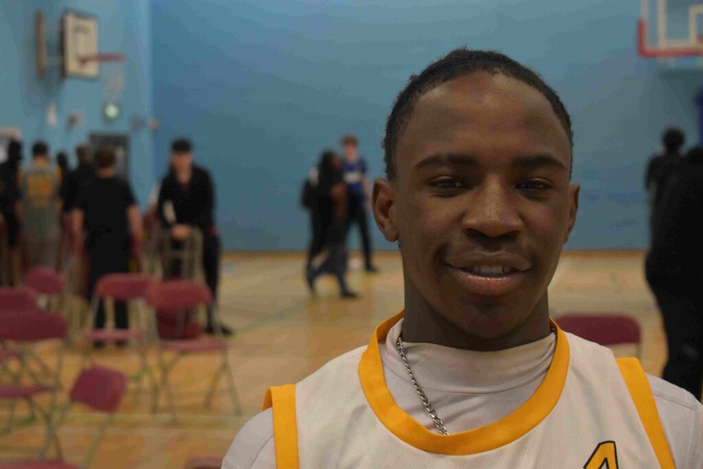 Young male student in sports uniform at Suffolk New College gymnasium, with other students and sports equipment in the background.