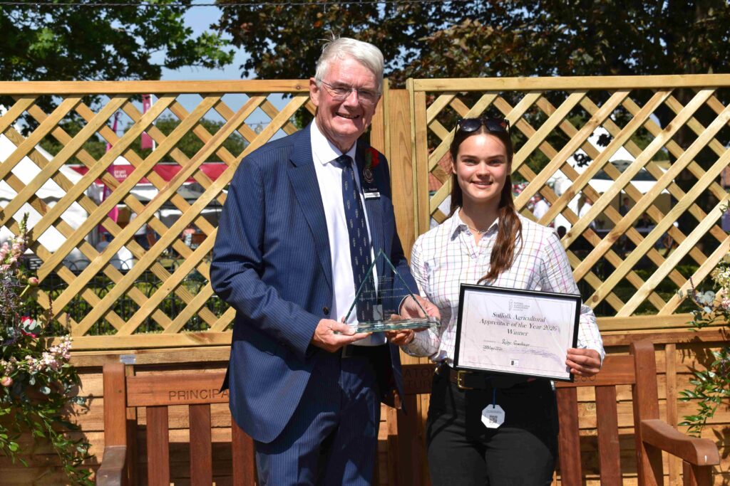 A man and a young woman standing outdoors in front of a wooden fence, with the man presenting an award and the woman holding a certificate, celebrating achievement at Suffolk New College.