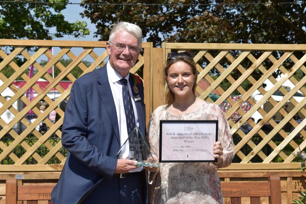 A young woman receiving an award from an older man at Suffolk New College, both smiling, outdoors with a wooden fence and trees in the background, celebrating achievement and success.