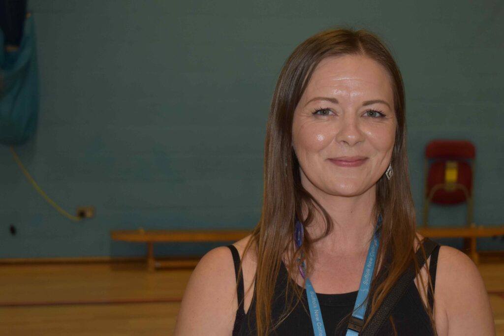 A friendly woman with long brown hair smiling at the camera, wearing a black top and lanyard, standing in a sports hall at Suffolk New College, promoting education and training.