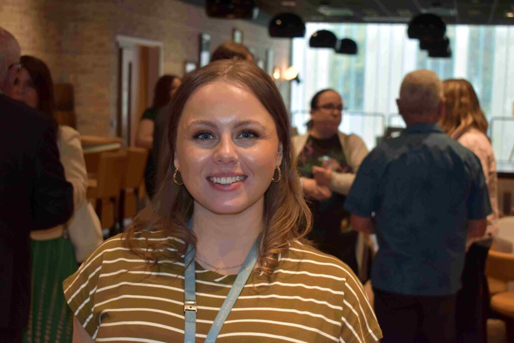 A smiling young woman with shoulder-length hair and a striped top, attending a Suffolk New College event with other students and staff in a modern, well-lit indoor setting.