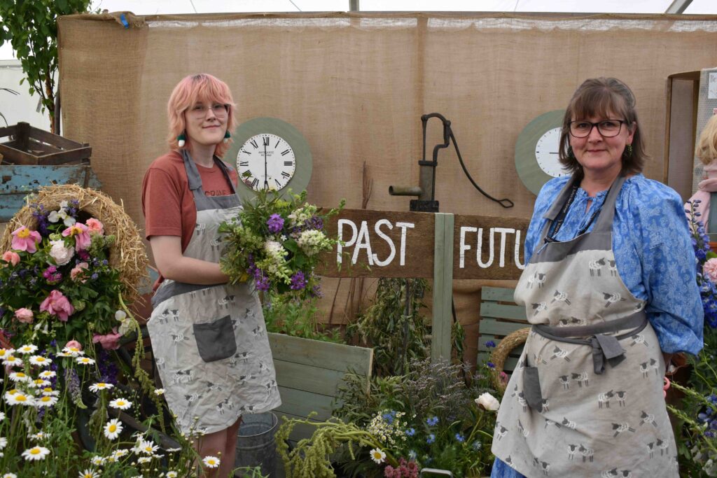 Two students at Suffolk New College standing in a garden display with flowers, plants, and a "Past" and "Future" sign, representing growth and progress in horticulture studies.