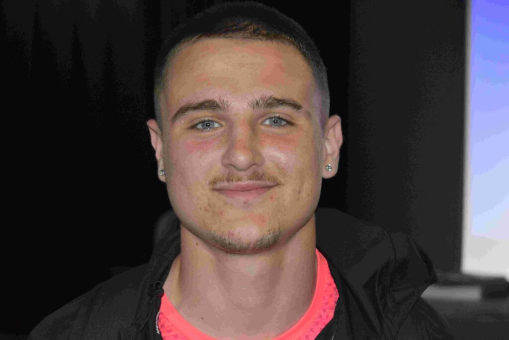 A young man with short dark hair and a slight smile, wearing a black jacket and pink shirt, standing indoors during a college visit at Suffolk New College.