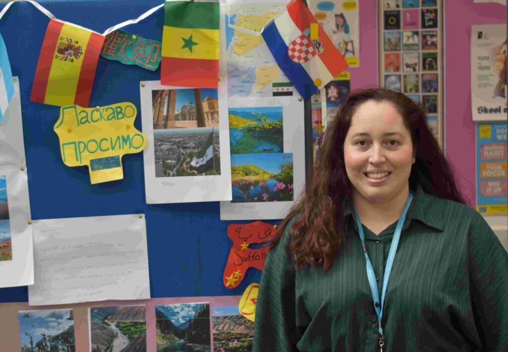 A young woman standing in front of a colourful display board with flags and travel photos at Suffolk New College, promoting international education and cultural diversity.