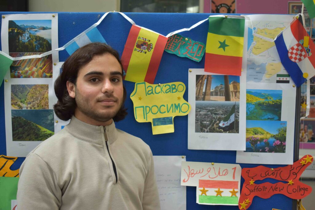 A young male student with long dark hair standing in front of a colourful display board decorated with flags, photos, and messages representing different countries at Suffolk New College.