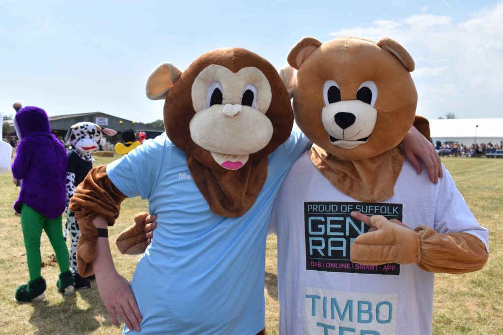 Two people dressed in monkey and bear mascot costumes at an outdoor event, promoting Suffolk New College with a crowd and tents in the background.