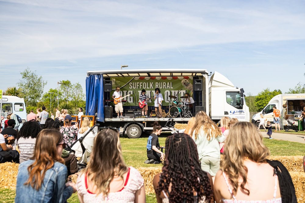 Outdoor community event at Suffolk New College featuring live music on a mobile stage, with students and local residents enjoying the event in a sunny, open field