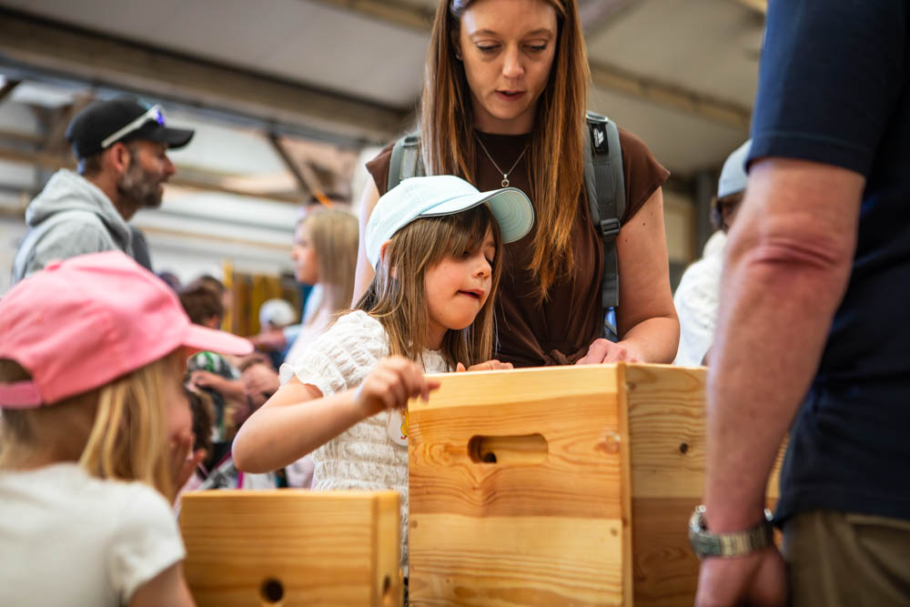 A young girl and her mother engaging with wooden craft activities at Suffolk New College's family fun day, surrounded by other families and outdoor event tents.
