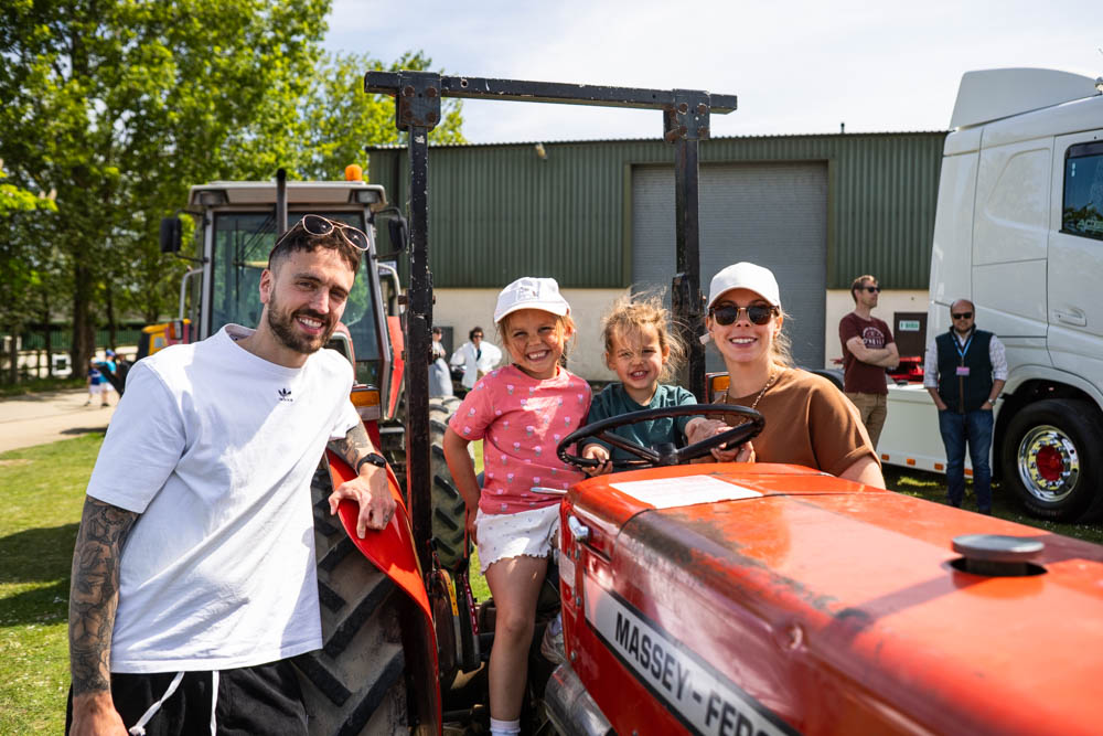 Smiling family riding a tractor at Suffolk New College's annual family fun day, with children and adults enjoying outdoor activities in sunny weather.