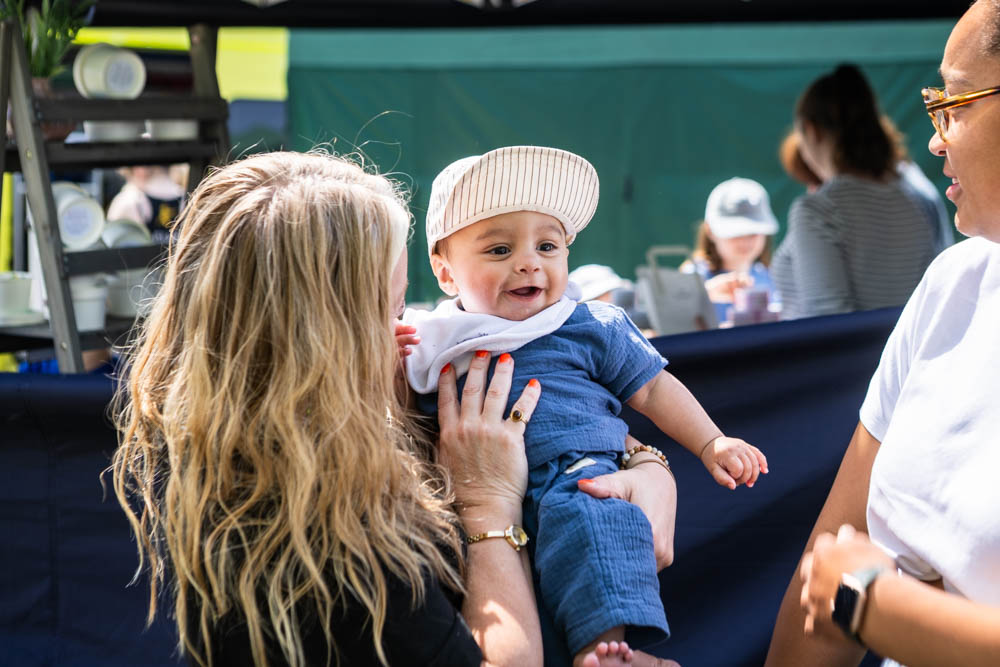 A woman holding a smiling baby boy at a family fun day event outdoors, with other families and children in the background, enjoying sunshine and activities.