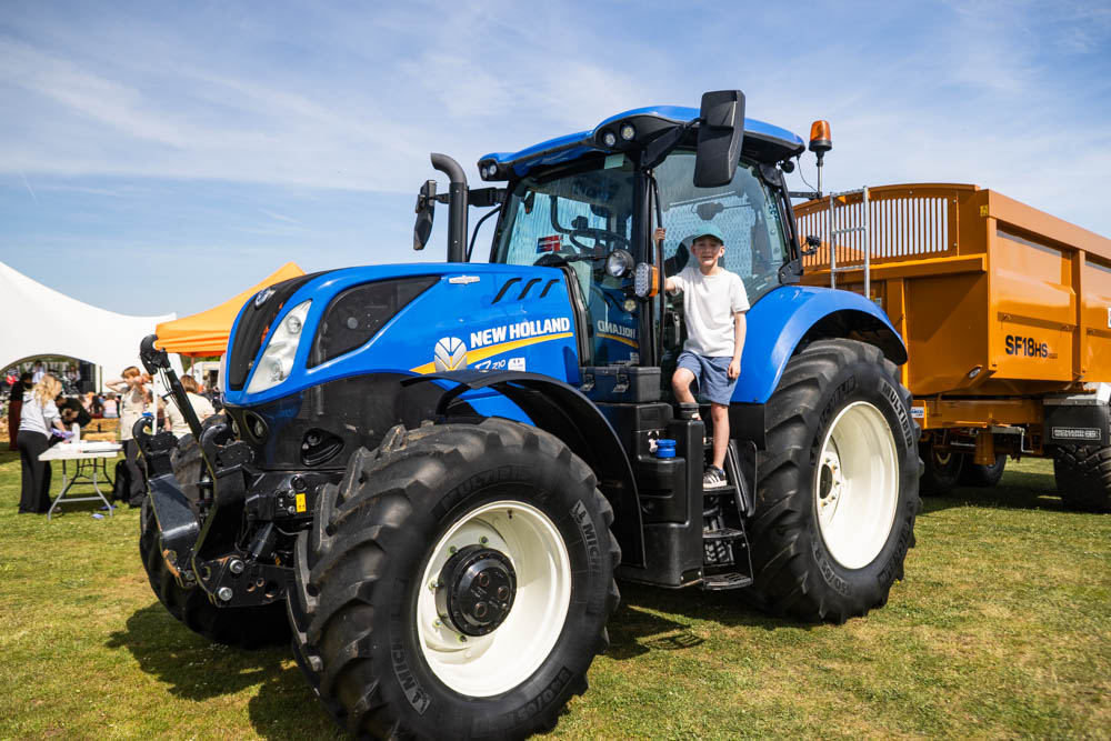 A young boy sitting on a large blue New Holland tractor at Suffolk New College during the annual family fun day, with a clear sky and other event activities in the background.