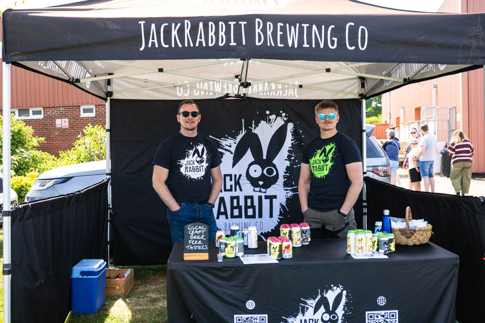 Two men standing behind a Jackrabbit Brewing Co stall at an outdoor event, with cans of beer and promotional materials, during a sunny family fun day.