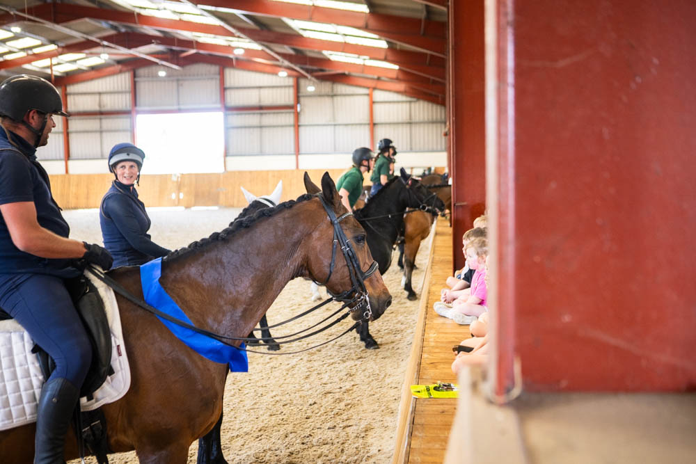 Students and instructors participating in a horse riding lesson at Suffolk New College's equestrian centre, with children watching from the sidelines during a family fun day.