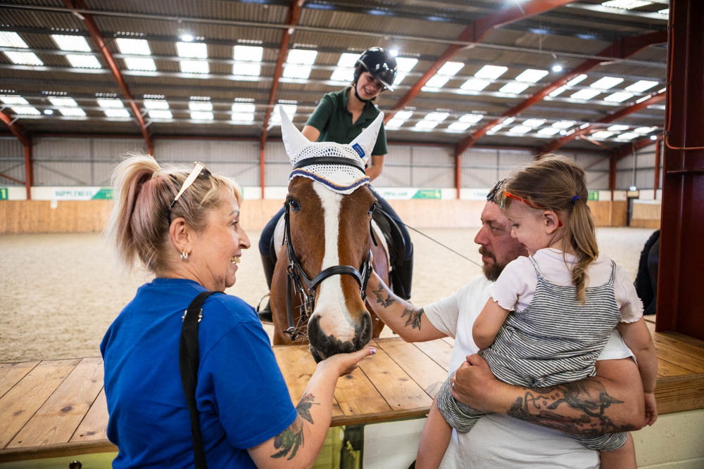 A young girl in riding gear interacts with a horse during a therapy session, while two adults, one with tattoos, support her at Suffolk New College's equestrian centre.