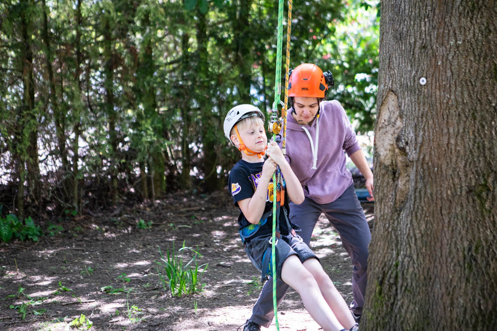 A young boy and an instructor enjoying tree climbing at Suffolk New College during a family fun day, surrounded by lush greenery and tall trees.