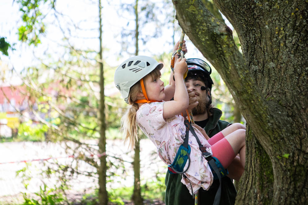A young girl wearing a helmet and harness enjoying a tree climbing activity with an instructor during a family fun day at Suffolk New College, surrounded by lush greenery.