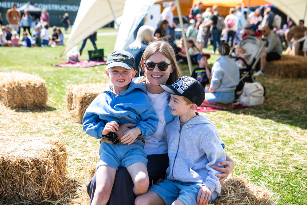Smiling woman with two young boys sitting on hay bales during a sunny outdoor family event at Suffolk New College, with tents and other families in the background.