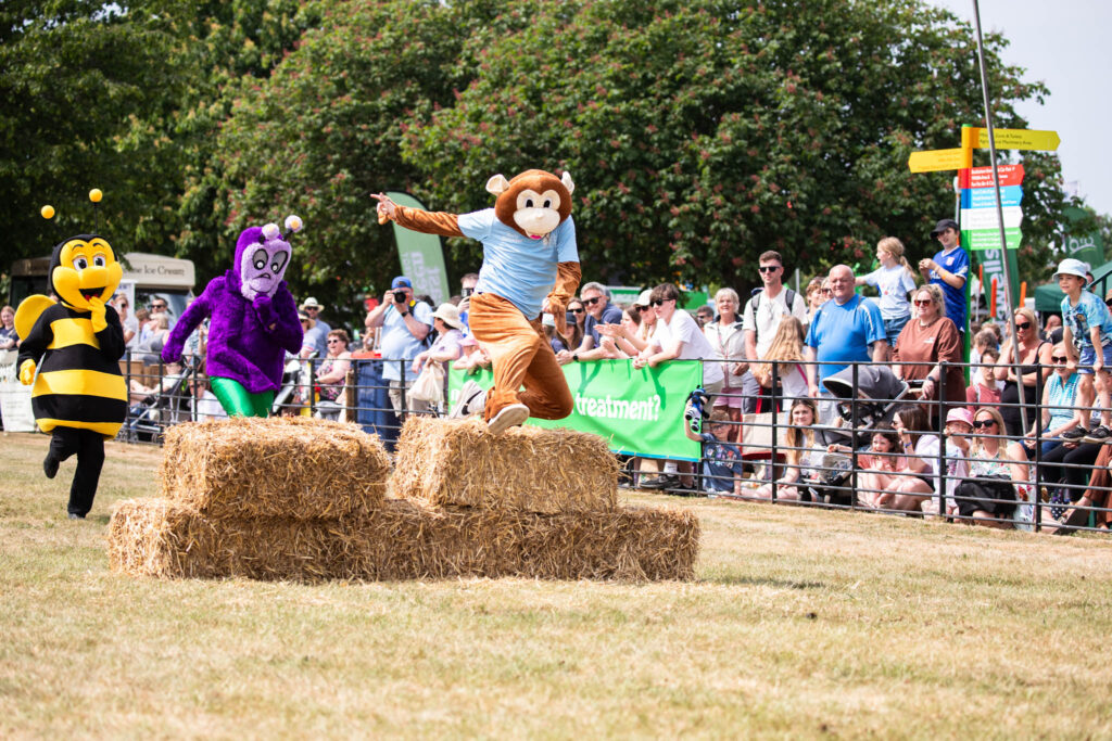 Costumed characters, including a monkey, bee, and purple creature, entertaining a crowd at Suffolk New College outdoor event with hay bales and spectators.
