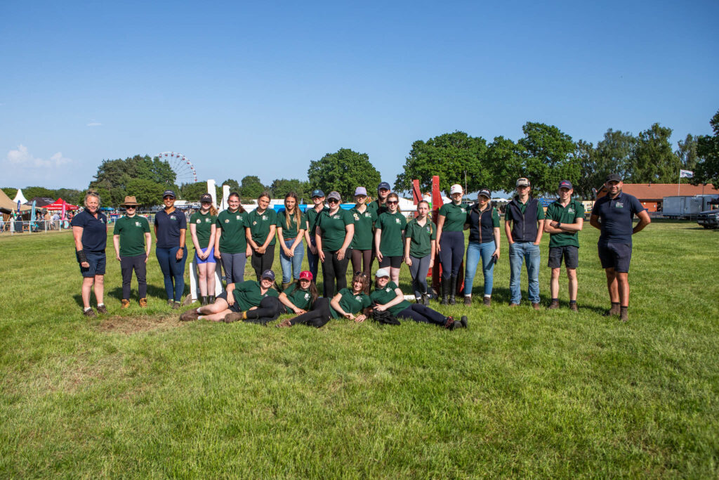 Group of students and staff from Suffolk New College standing on a grassy field during an outdoor event, with clear blue sky and amusement park rides in the background.