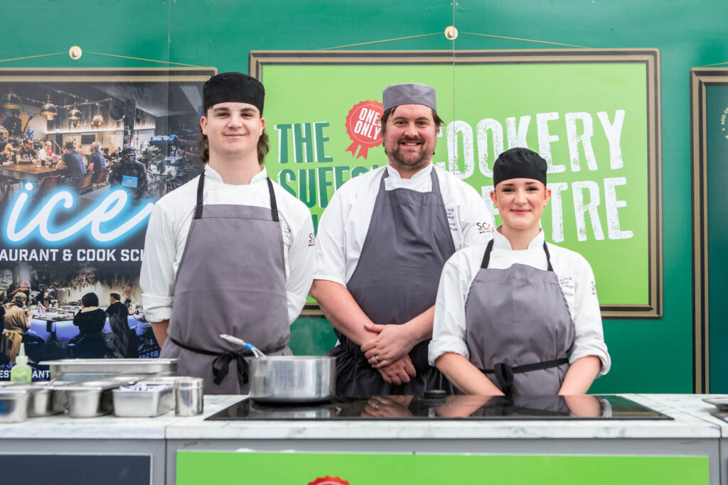Three Suffolk New College students in chef uniforms and aprons standing behind a cooking station, smiling at the camera during a culinary demonstration at the Suffolk Food Discovery Centre.