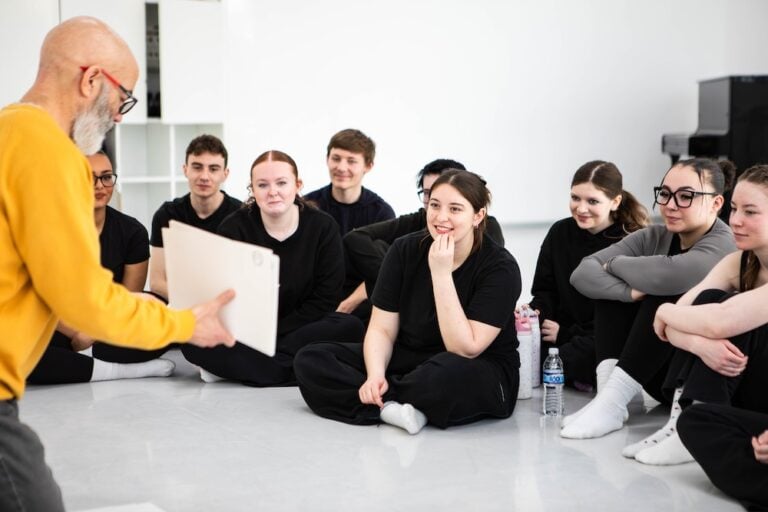 College students sitting on the floor listening attentively to a teacher during a workshop at Suffolk New College, showcasing student engagement and learning environment.