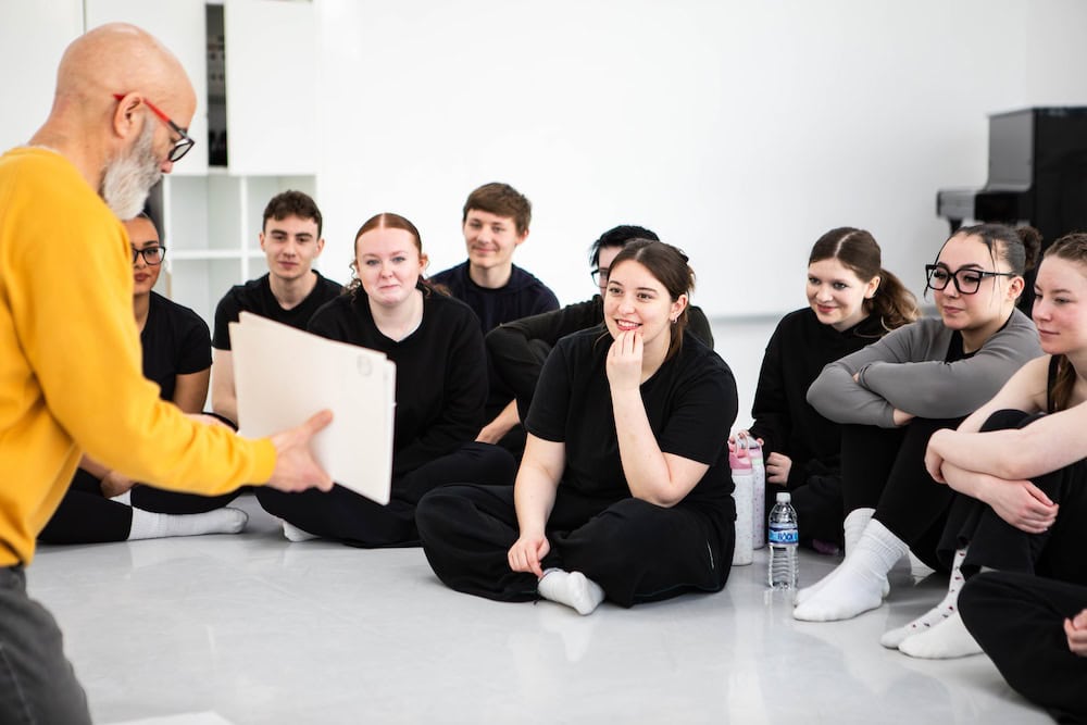 College students sitting on the floor listening attentively to a teacher during a workshop at Suffolk New College, showcasing student engagement and learning environment.