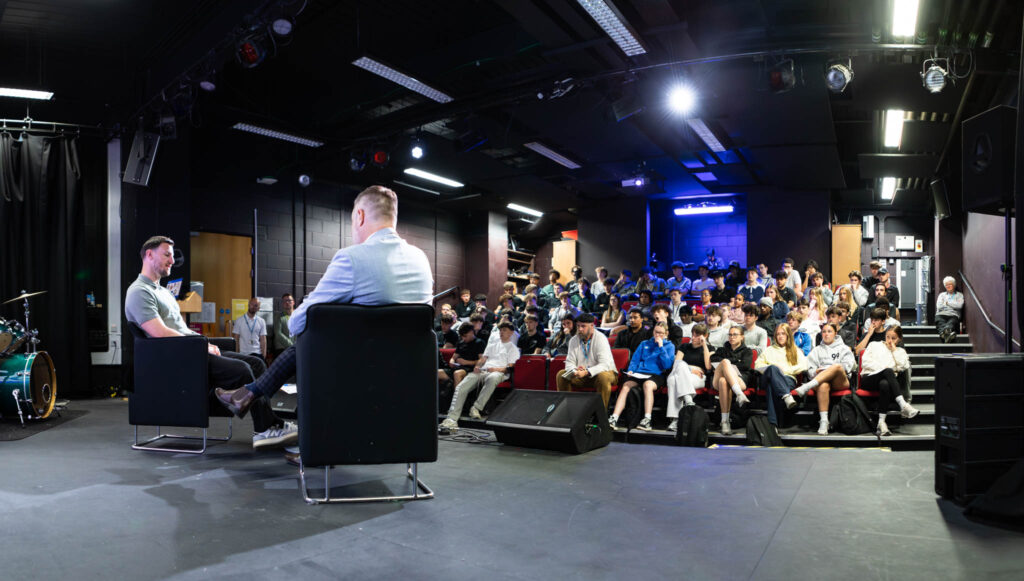 A large audience attending a lecture at Suffolk New College, with two speakers seated on stage engaging with students in a modern, well-lit auditorium.