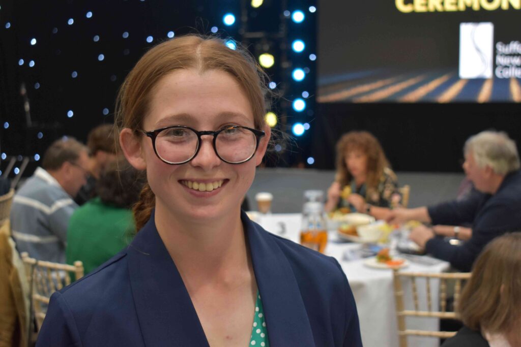 Young woman with glasses smiling at Suffolk New College awards ceremony, with other attendees and a stage in the background.