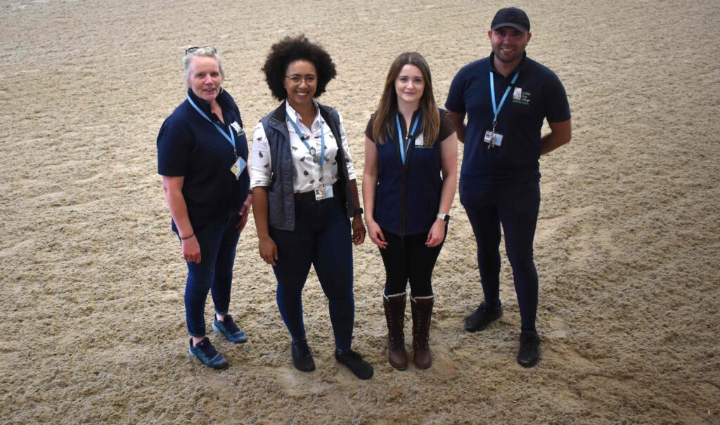 Group of four students and staff at Suffolk New College equine centre, celebrating 20 years of the facility, standing on sandy riding arena.