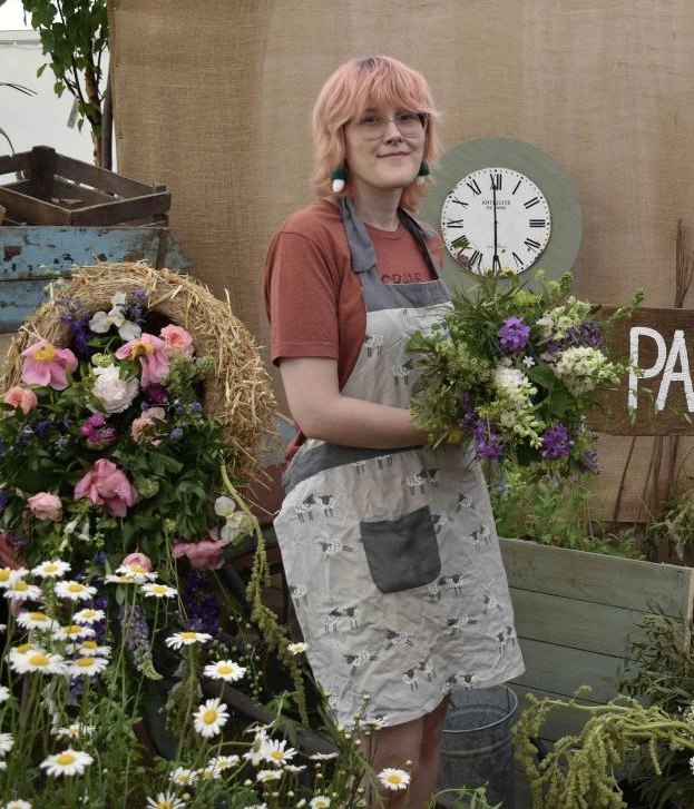 A college student with pink hair and glasses holding a bouquet of flowers at the Suffolk New College showcase event, surrounded by floral displays and garden decorations.