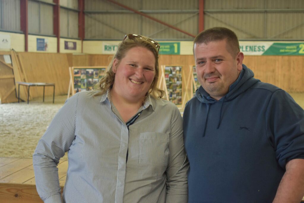 Two smiling individuals at Suffolk New College equine centre, celebrating 20 years of the facility's opening, with horse riding arena in background.