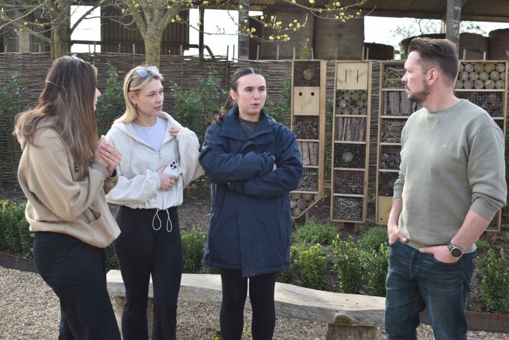 Group of students and instructor engaging in an outdoor learning activity at Suffolk New College, with a garden and educational structures in the background.