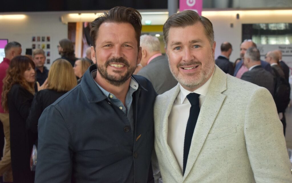 Two men smiling at a Suffolk New College event, dressed professionally, with a crowd of people in the background, showcasing college community and engagement.