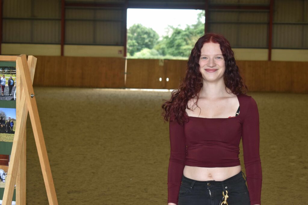 A smiling female student with curly red hair standing inside an indoor riding arena at Suffolk New College, celebrating the college's equine centre anniversary.
