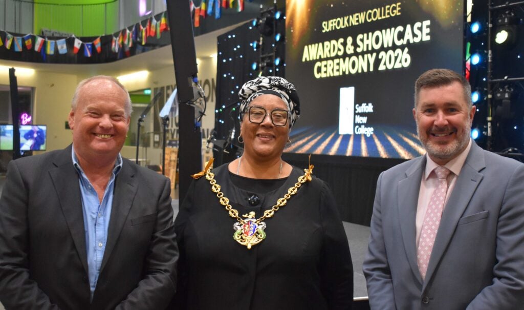Three individuals at Suffolk New College Awards & Showcase Ceremony 2026, celebrating academic achievements in a festive setting with a decorated stage and a large screen.