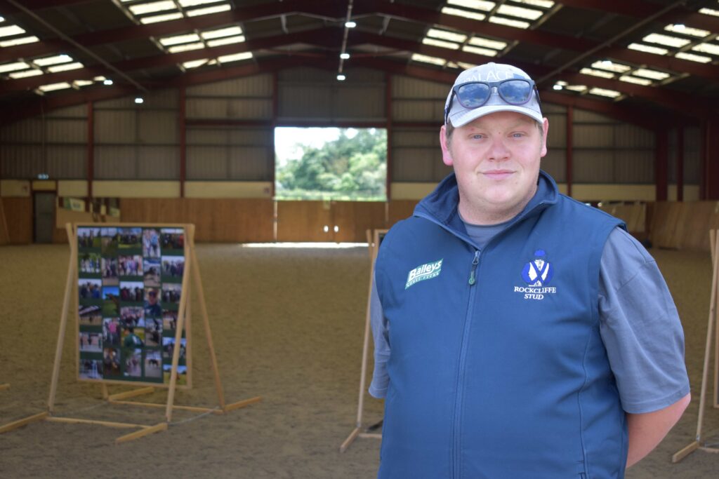 Young man in Suffolk New College equine centre wearing a blue vest and cap, with a photo display in the background, celebrating 20 years of the equine facility.