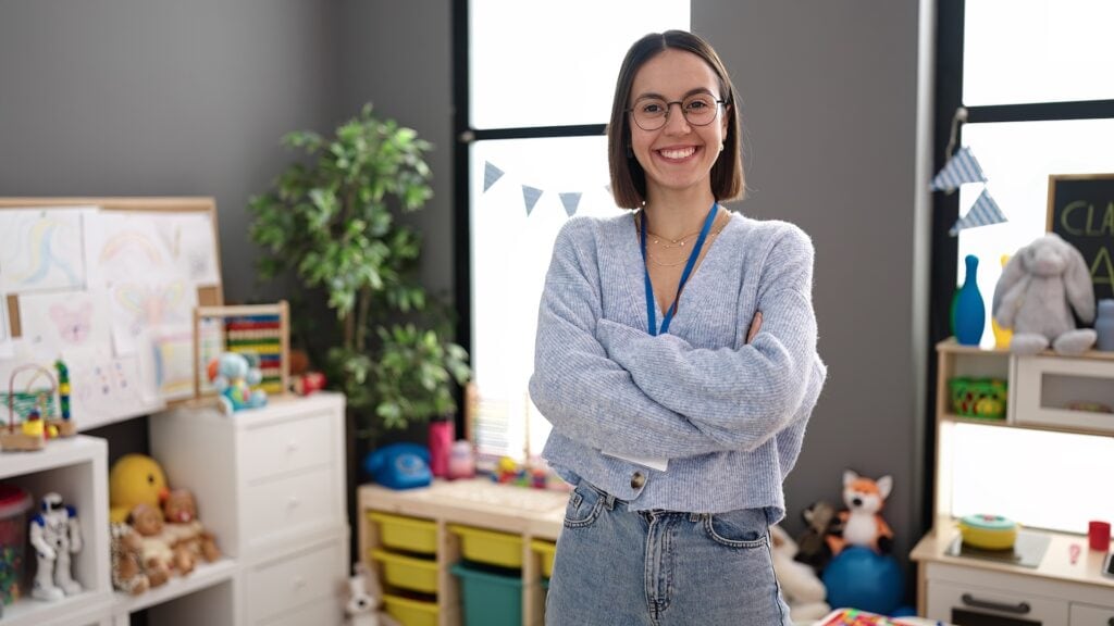 A smiling young woman with glasses and a grey sweater standing confidently in a classroom filled with educational toys, books, and colourful decorations at Suffolk New College.