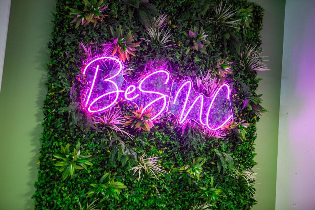 Vibrant pink neon sign reading 'Be SMC' mounted on a lush green wall with various plants at Suffolk New College, promoting student engagement and college spirit.