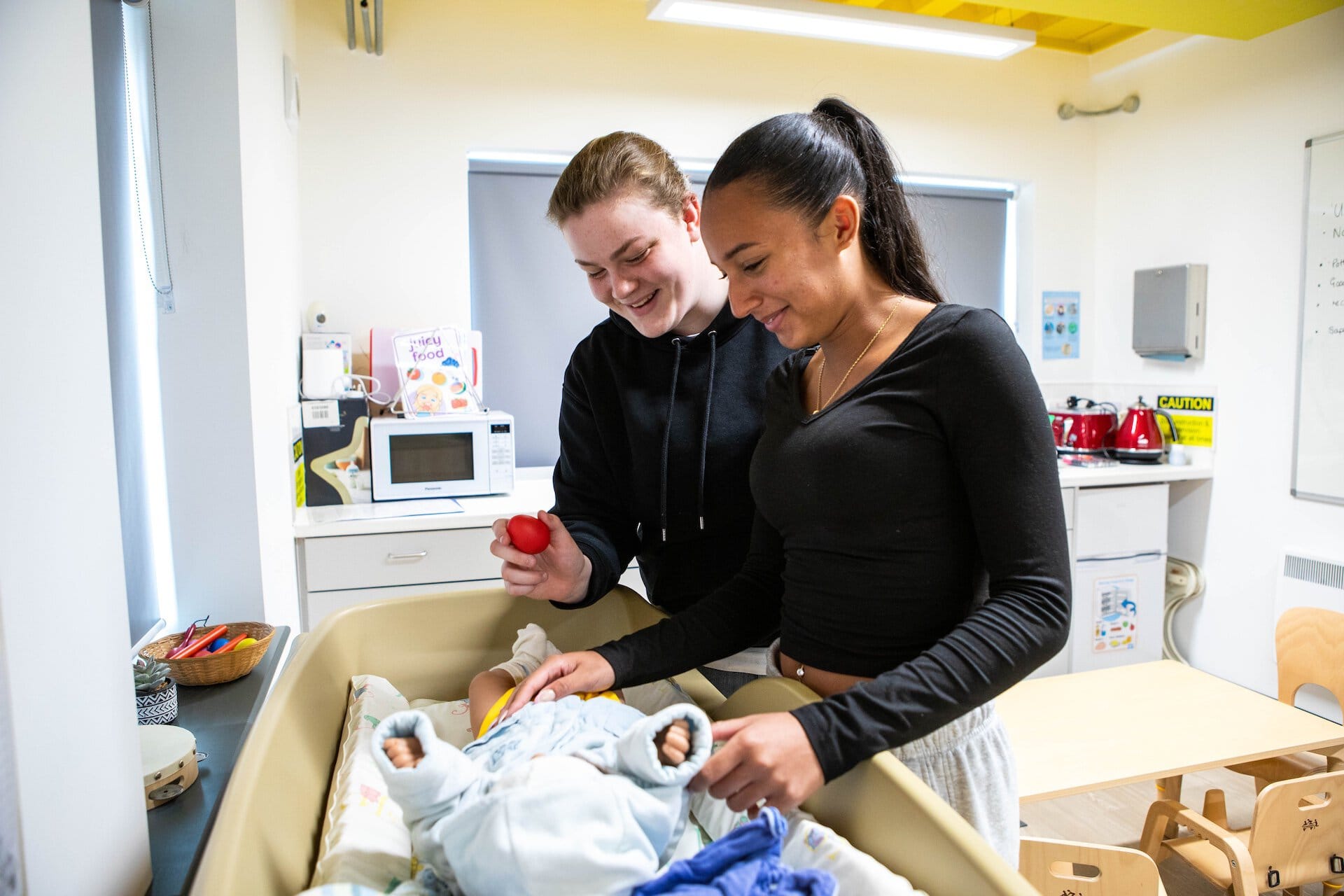 Childcare students at Suffolk New College demonstrating infant care techniques with a baby doll in a simulated nursery environment.