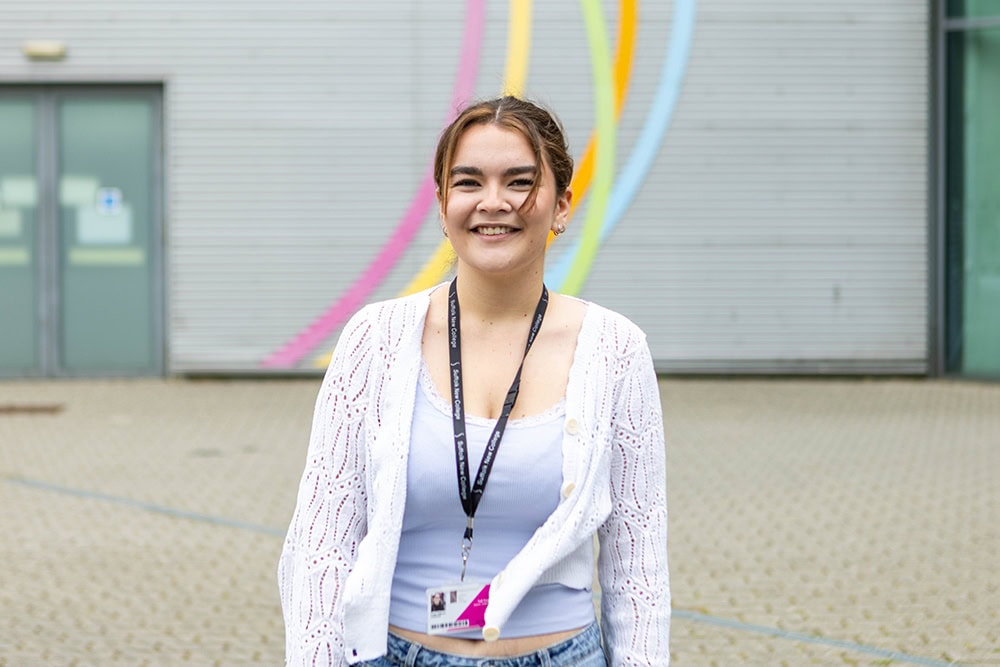 Smiling young woman with a lanyard standing outside Suffolk New College, with colourful wall art in the background, representing education and student life.
